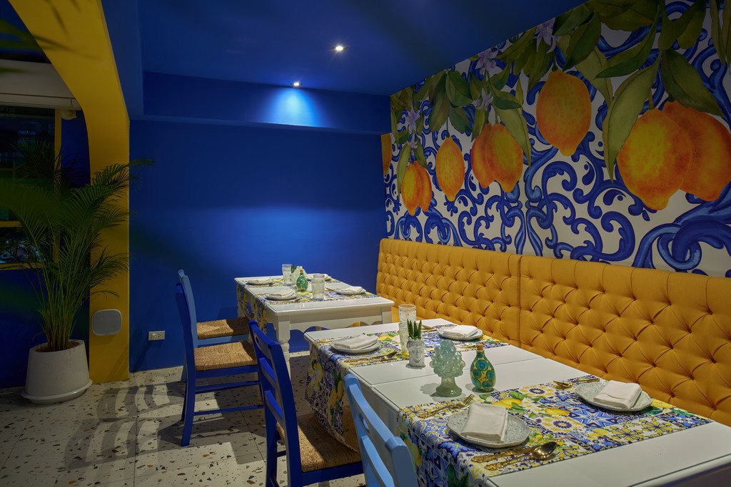 Yellow tufted banquette under a hand painted lemon mural, with blue chairs and tables set with Sicilian ceramics inside Mandorla, Chiang Mai